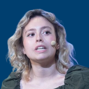 Elsa Arnáiz Chico hablando con micrófono de solapa, vestida con blusa verde, frente a un fondo azul.