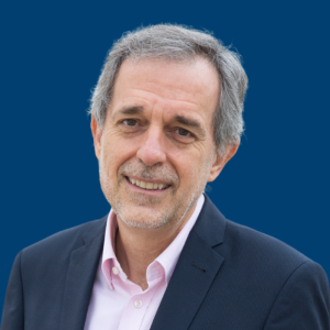 Juan Fernández Palacios vestido con traje oscuro y camisa rosada, frente a un fondo azul.