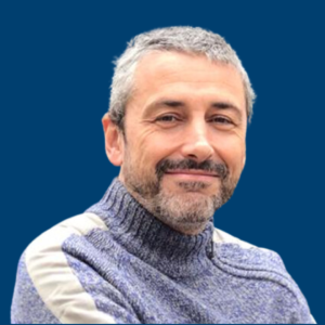 Mariano Sánchez sonriendo, vestido con un jersey gris frente a un fondo azul.