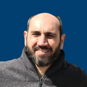 Pedro Galatas sonriente con chaqueta oscura posando frente a un fondo azul.